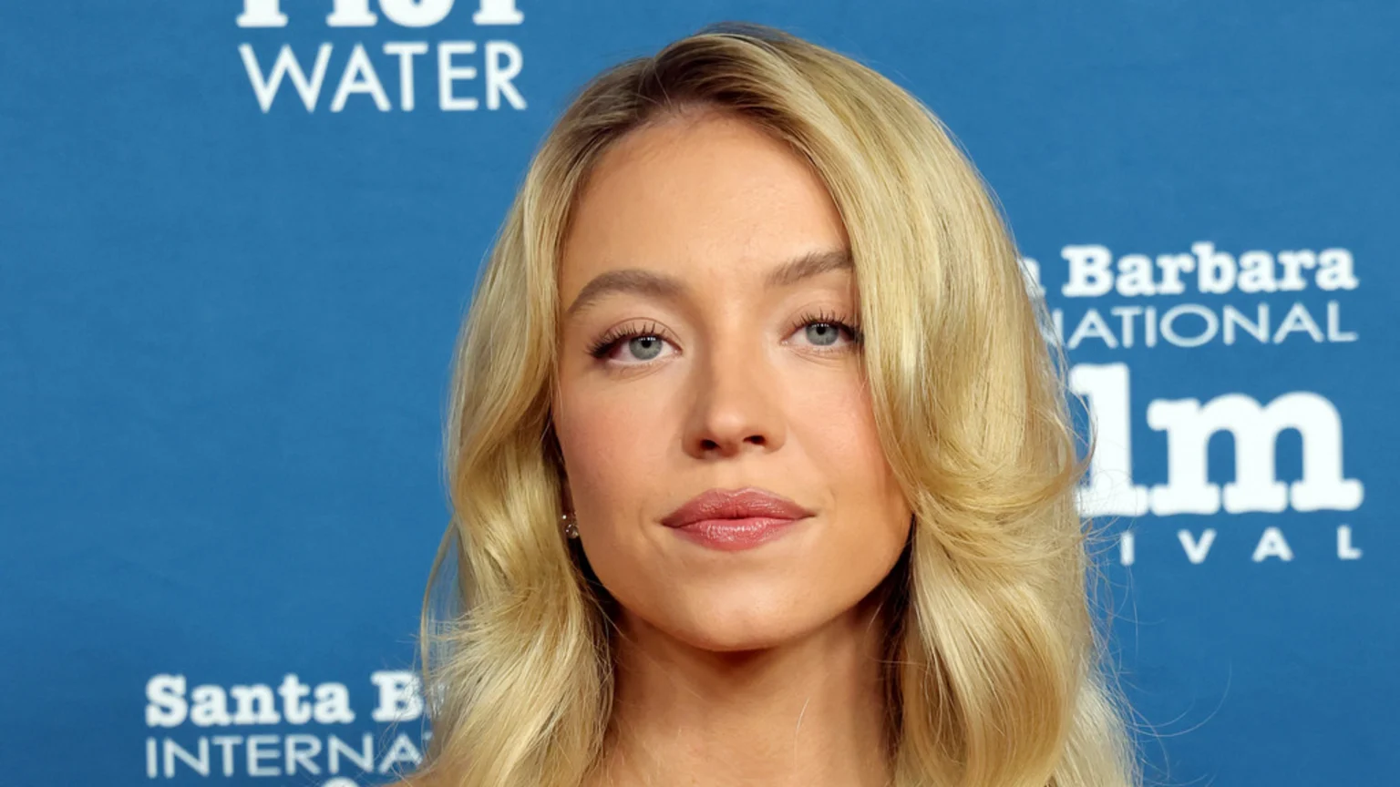 A close-up headshot of actress Sydney Sweeney with blonde hair and light eyes, wearing natural makeup, against a blue backdrop with partial white text from an event.