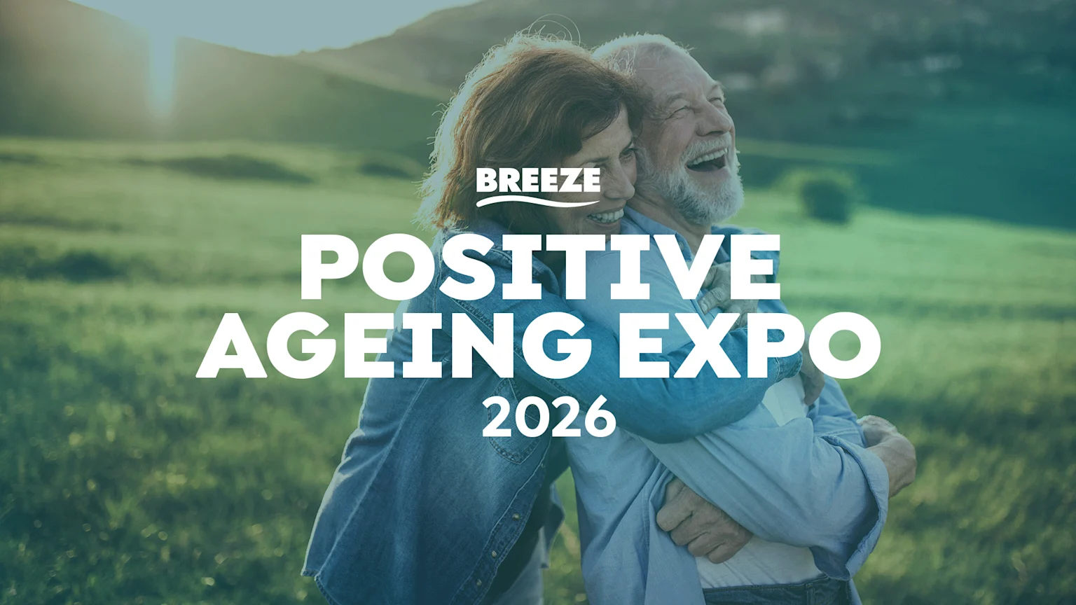 A man and a woman, both older adults, are embracing and laughing joyfully in a sunny, green field with hills, overlaid with text "BREEZE POSITIVE AGEING EXPO 2026".