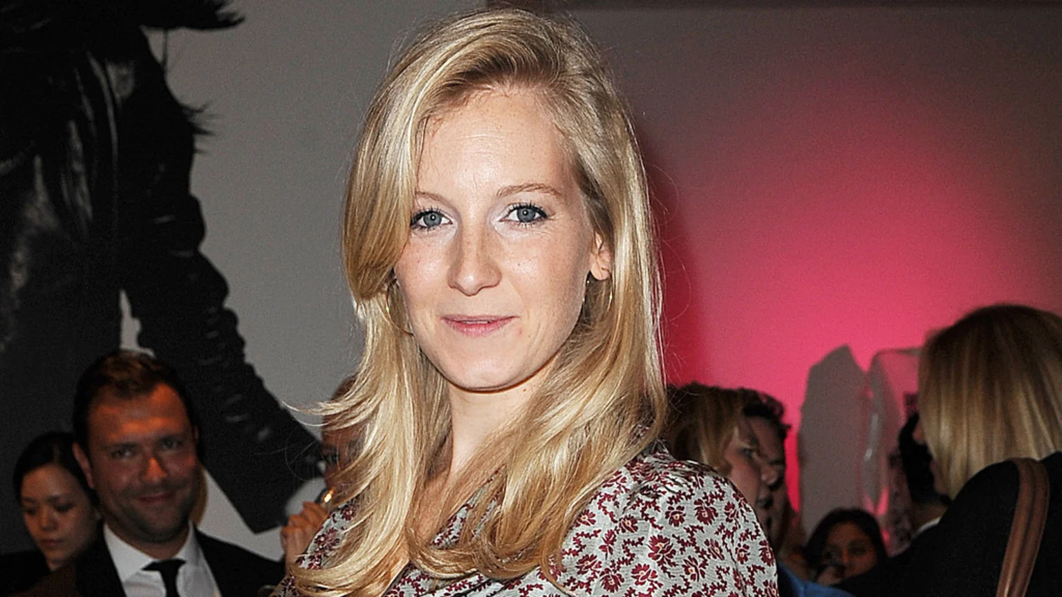 A blonde woman with blue eyes, wearing a patterned top, smiles slightly at the camera in a crowded room with a pink-lit wall.