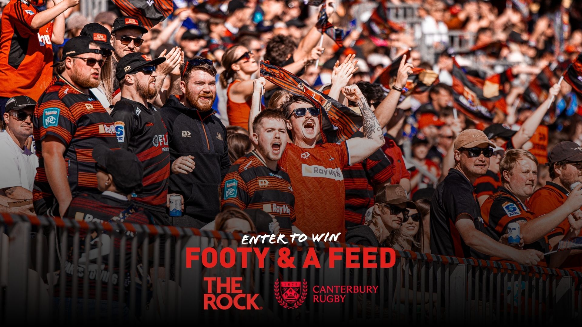 A cheering crowd of excited rugby fans, many in black and orange team jerseys, raising their arms and celebrating at a sunny outdoor game.