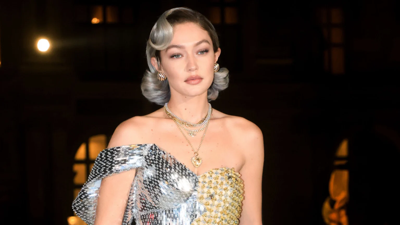 Gigi Hadid poses in a strapless dress, half silver sequins and half gold, with wavy grey hair, multiple necklaces, and gold hoop earrings against a dark background.