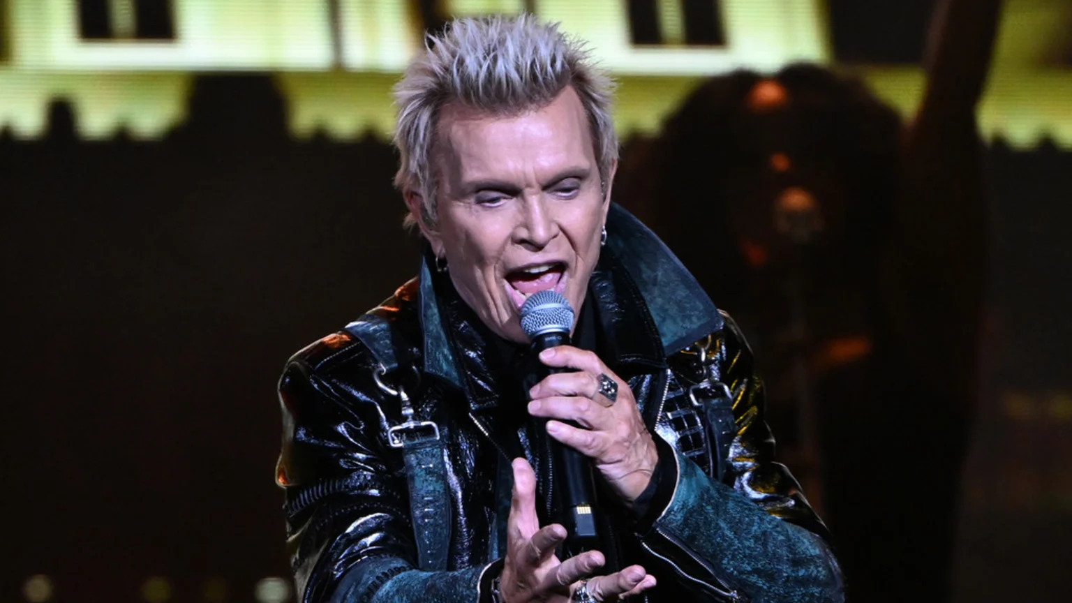 Billy Idol, a man with spiky blonde hair, sings into a microphone, wearing a shiny black and dark teal jacket, with his mouth open and rings on his fingers on a dark stage.