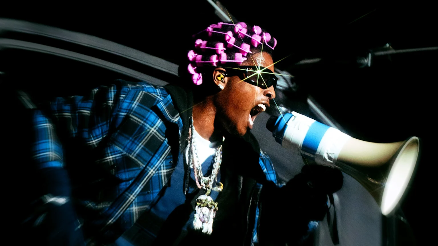 A$AP Rocky, wearing pink hair rollers, sunglasses with a starburst flare, a blue plaid shirt, and chains, shouts into a megaphone.