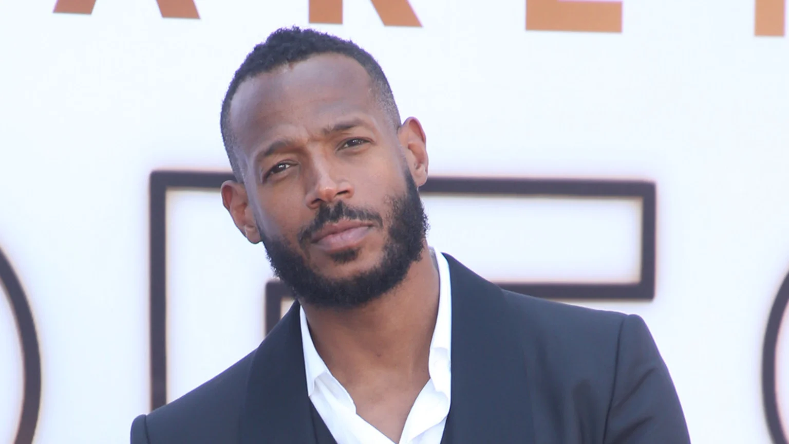 Marlon Wayans, a man with a beard and short dark hair, wearing a dark suit and white shirt, looks intently at the camera against a blurry light background.