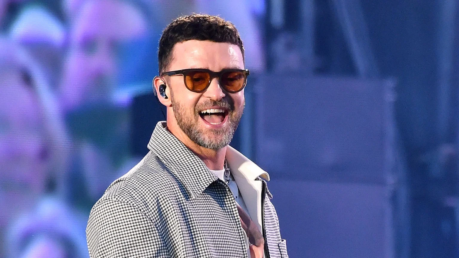 Justin Timberlake smiles broadly, wearing dark sunglasses with orange lenses, a black and white gingham shirt, and an in-ear monitor, against a blurred purple and blue background.