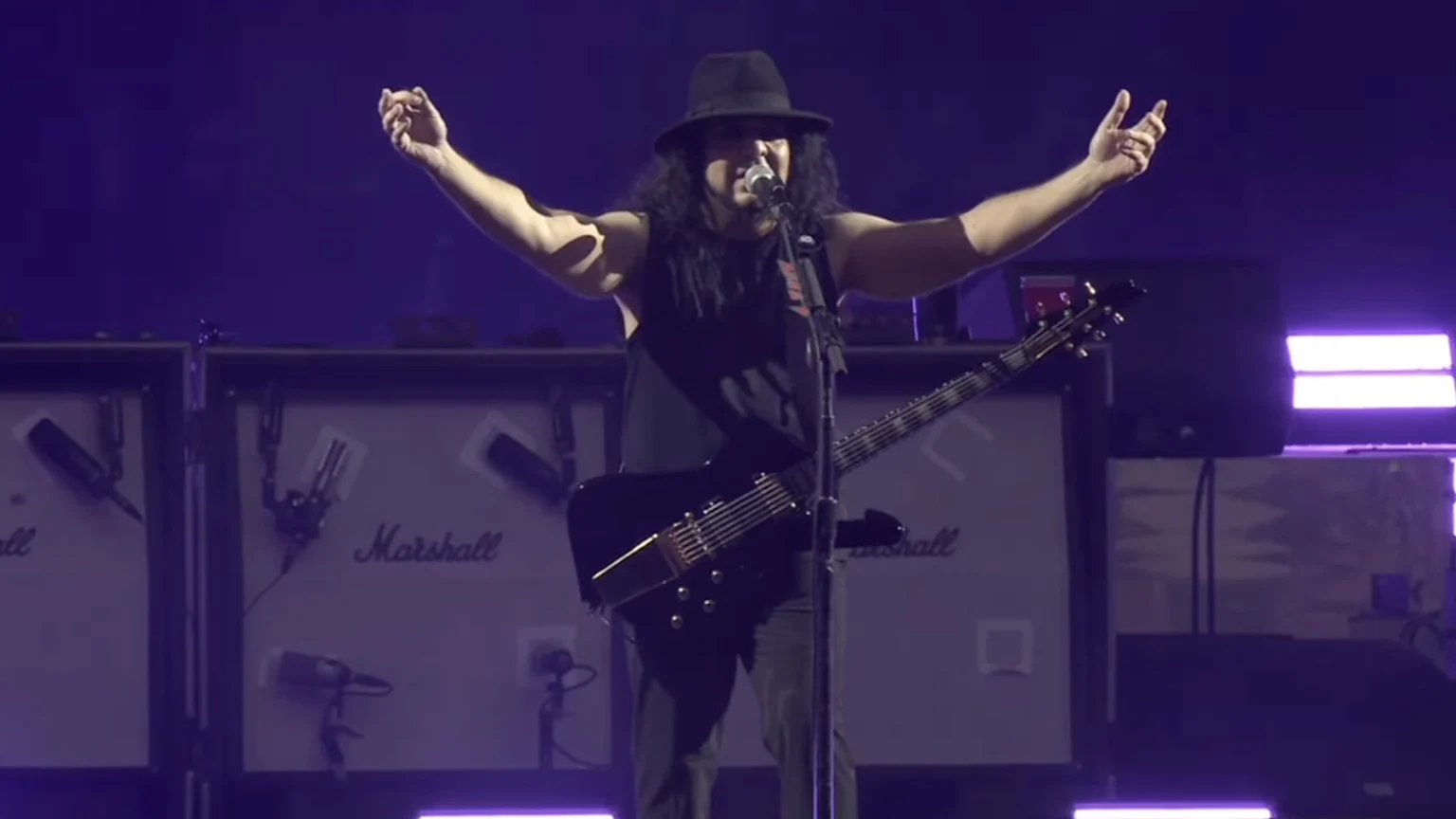 A male musician with long dark hair, wearing a black hat and tank top, stands on a dark stage with arms raised, holding a black electric guitar in front of several Marshall amplifiers.