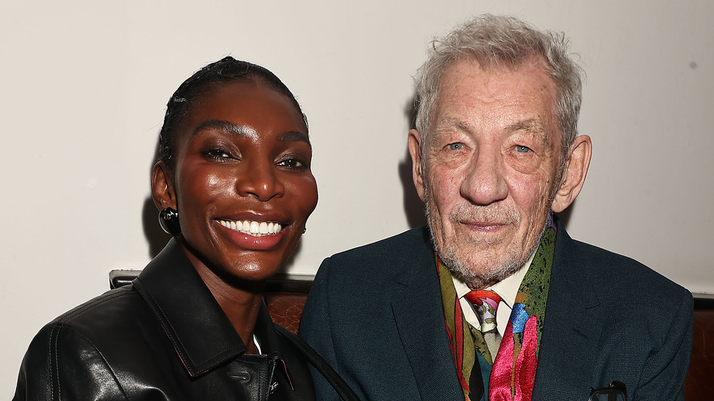 Michaela Coel, a smiling Black woman in a black leather jacket, poses next to an older white man with gray hair, a beard, and a colorful scarf.
