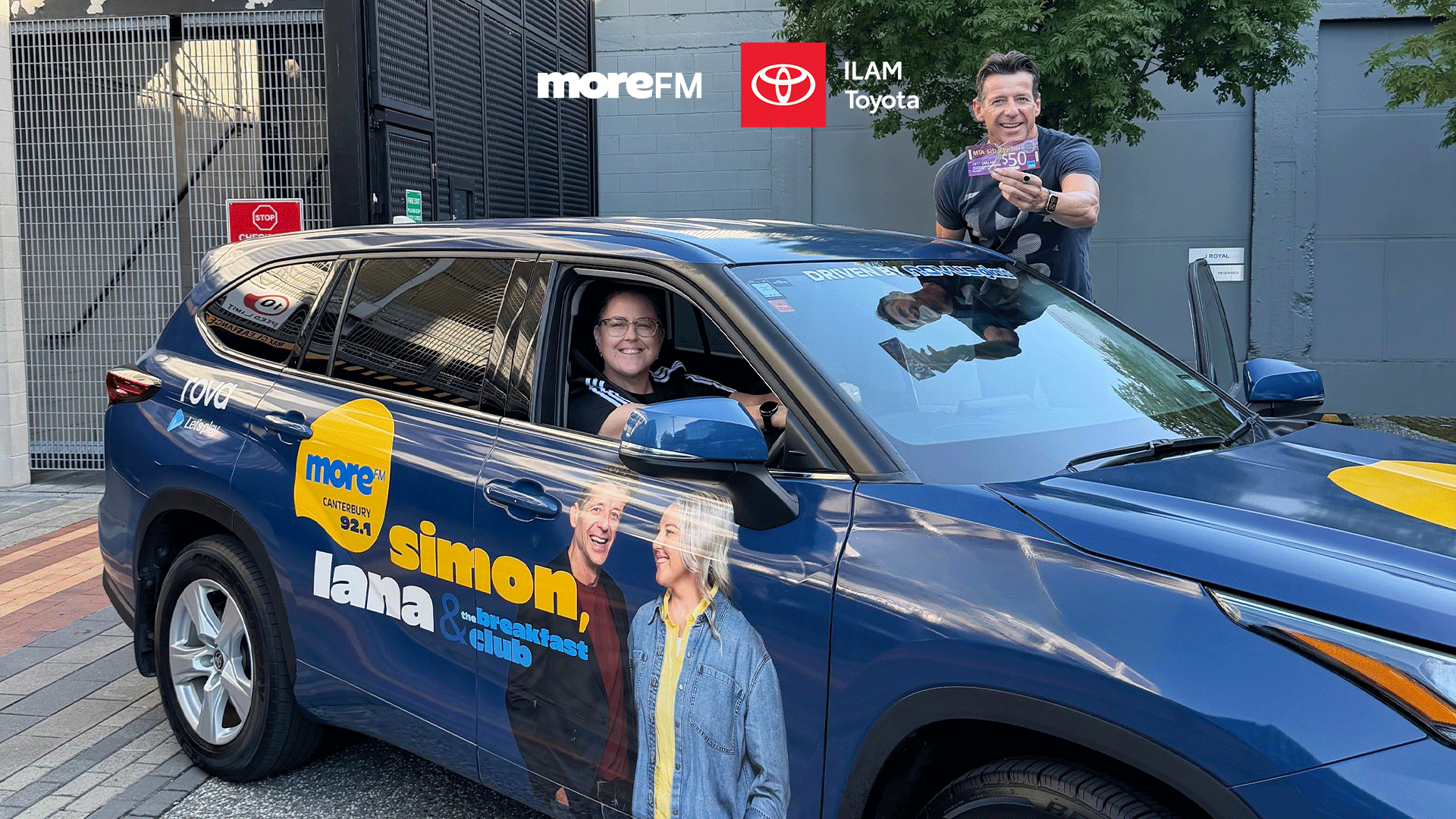 A blue ILAM Toyota SUV, branded with moreFM logos and "Simon, Lana & The Breakfast Club" graphics, with a woman in the driver's seat and a man holding a $50 gift card.