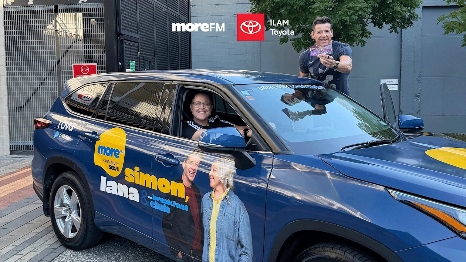 A blue ILAM Toyota SUV, branded with moreFM logos and "Simon, Lana & The Breakfast Club" graphics, with a woman in the driver's seat and a man holding a $50 gift card.