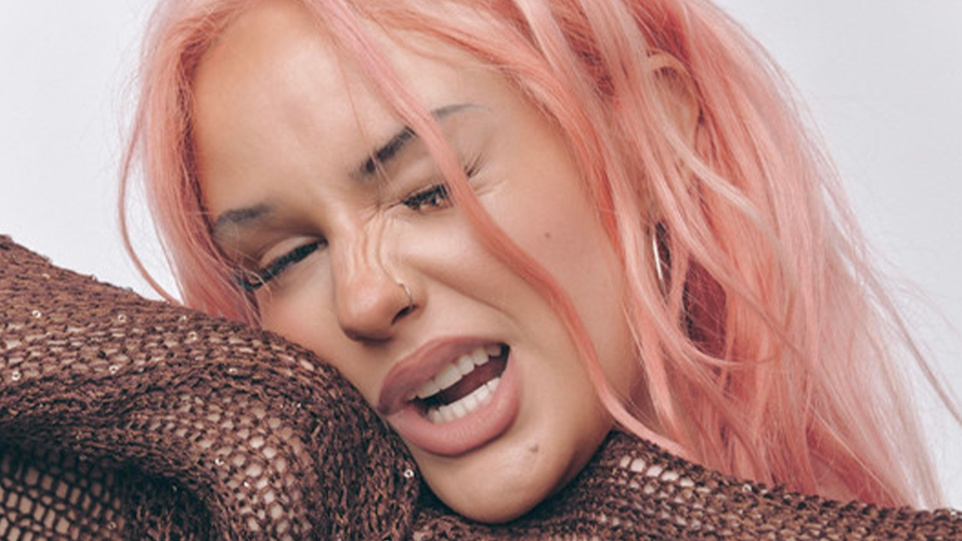 A person with pink hair and a nose ring is grimacing with a wrinkled nose, squinted eyes, and open mouth, wearing a brown textured top.