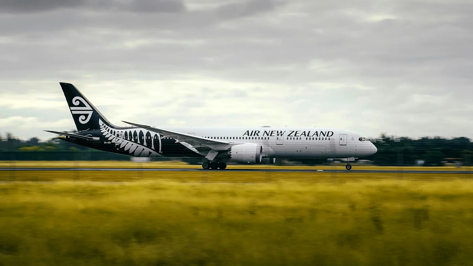 An Air New Zealand Boeing 787 Dreamliner aircraft, featuring a black tail with a white Koru and fern livery, is in motion on a runway with blurred green-yellow grass in the foreground under a cloudy sky.
