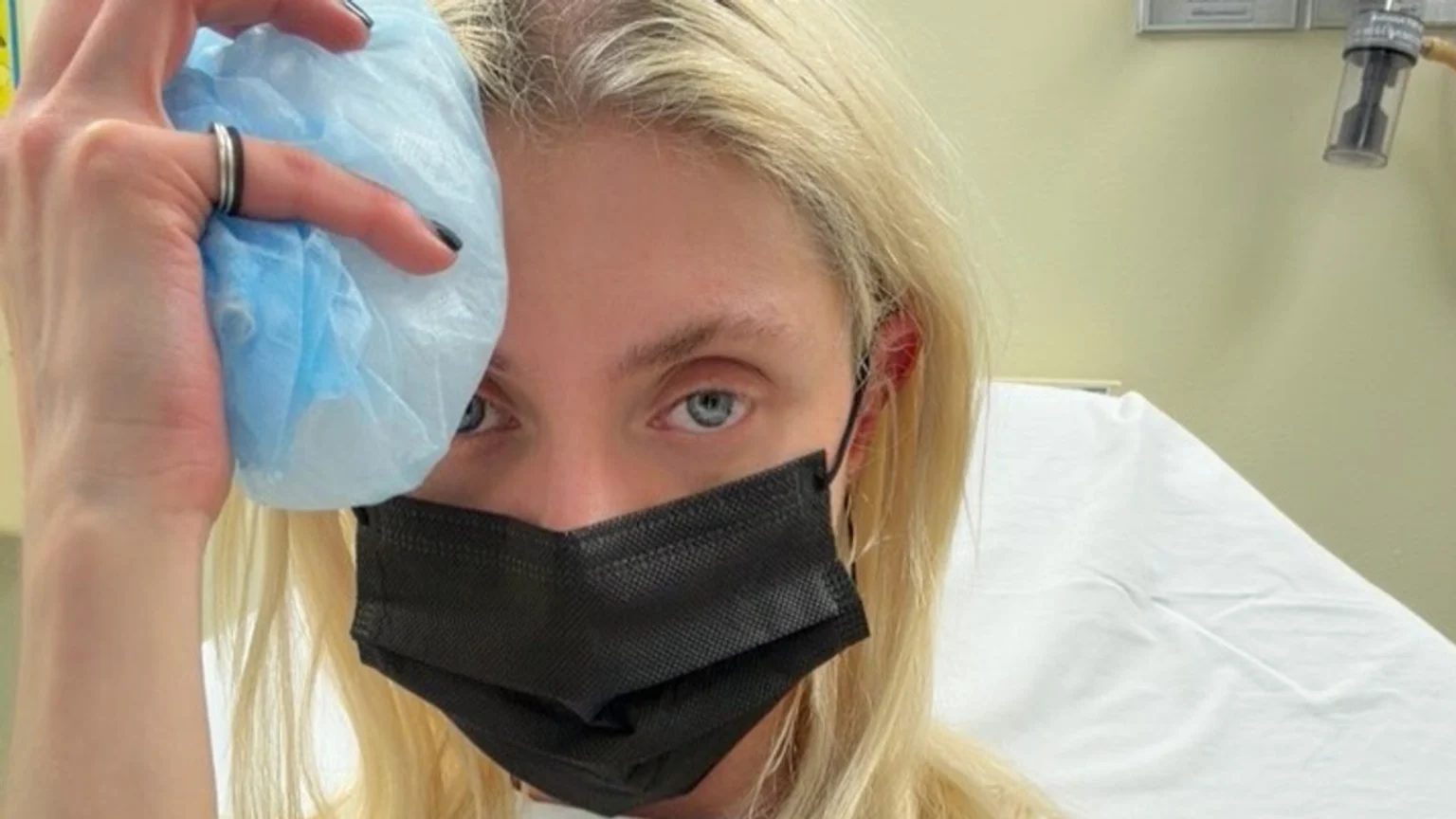 A blonde person wearing a black mask holds a blue ice pack to their forehead while lying in a hospital bed, looking at the camera.
