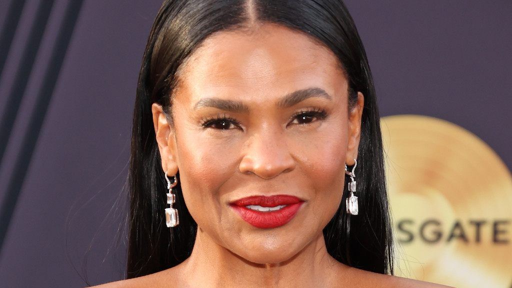 A close-up portrait of a smiling woman with dark hair, dark eyes, and red lipstick, wearing silver rectangular drop earrings against a blurred purple and gold background.