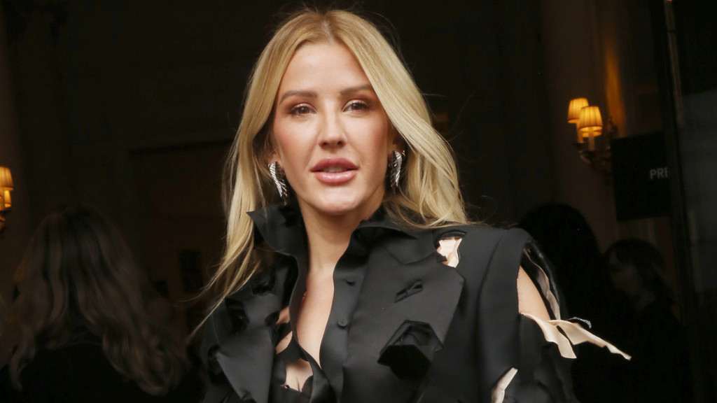 Ellie Goulding, with long blonde hair, wears prominent silver wing earrings and a black top with textured, torn fabric details.