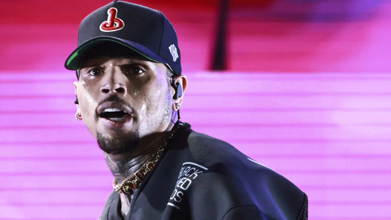 Chris Brown with a sweaty face and open mouth, wearing a black baseball cap with a red logo, a gold chain, and an in-ear monitor, against a vibrant pink, horizontally lined background.