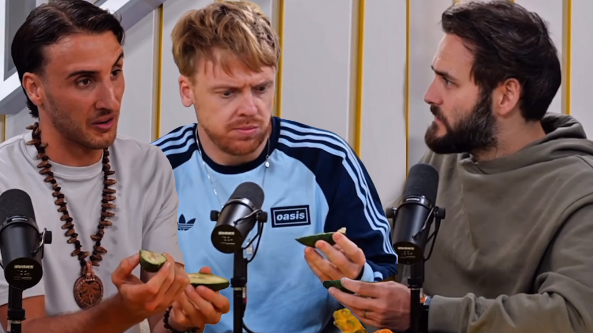 Three men sit at a table with microphones, holding green fruit slices; the middle man appears surprised as the others look on.
