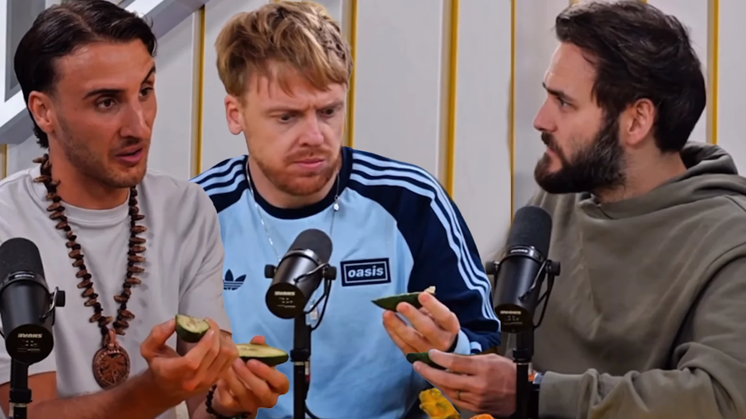 Three men sit at a table with microphones, holding green fruit slices; the middle man appears surprised as the others look on.