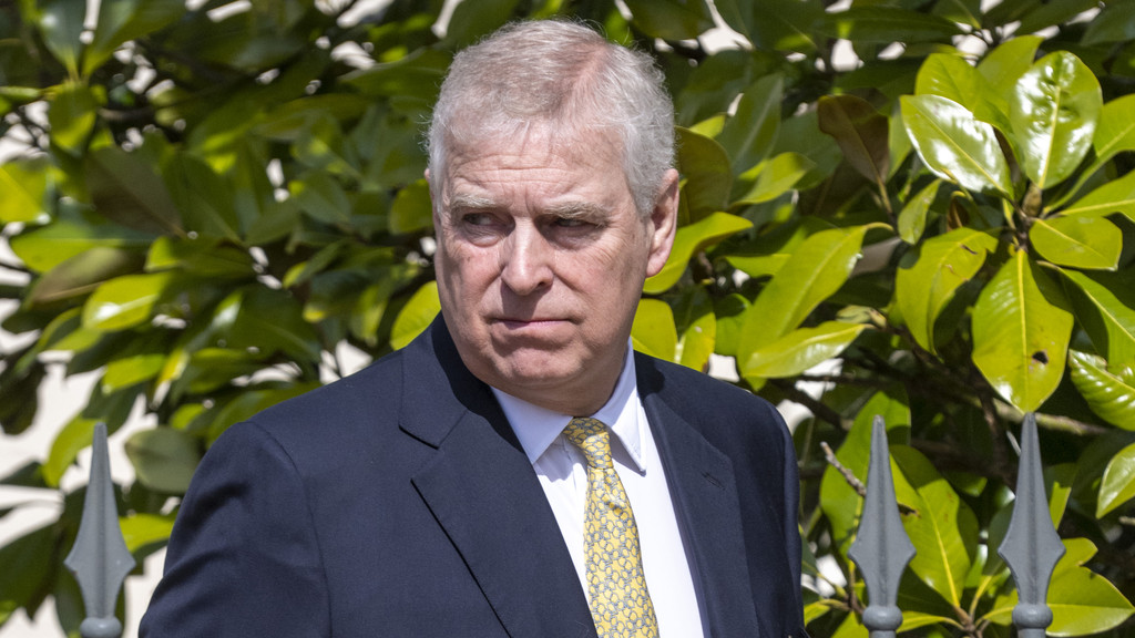 Prince Andrew, a man with grey hair, wears a dark suit, white shirt, and yellow patterned tie, looking serious, with lush green leaves and a grey spiked metal fence behind him.