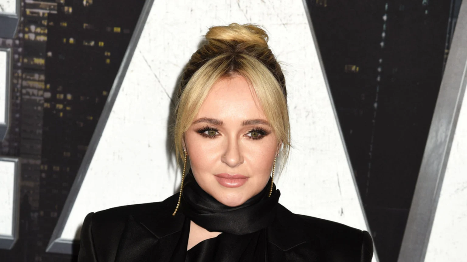 Hayden Panettiere looks directly at the camera, wearing a black blazer and scarf, with her blonde hair styled in a high bun and bangs, and long gold beaded earrings.