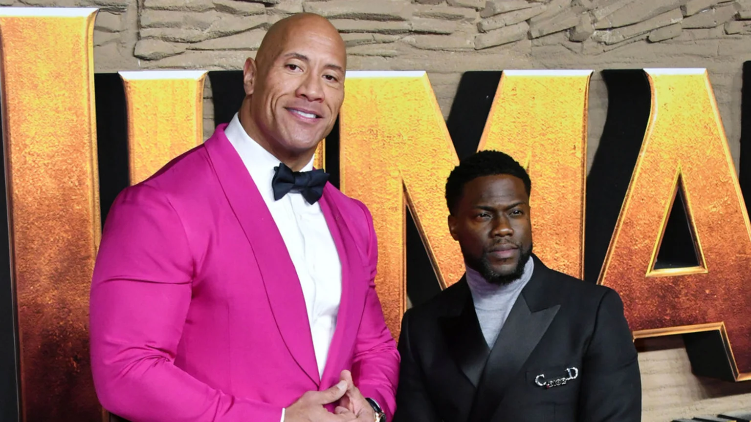 Dwayne "The Rock" Johnson in a pink suit smiles next to Kevin Hart in a black suit and turtleneck, both in front of a golden "JUMANJI" sign.