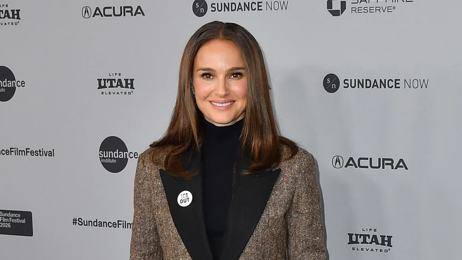 Natalie Portman smiles at the camera, wearing a brown tweed blazer with black lapels over a black turtleneck, and an "ICE OUT" pin, in front of a logo backdrop.