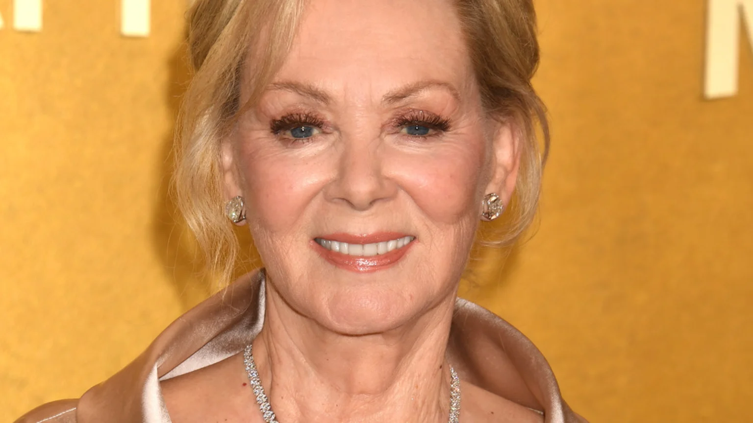 Close-up of Jean Smart smiling, wearing diamond earrings and a necklace, with blonde hair and blue eyes, against a gold background.