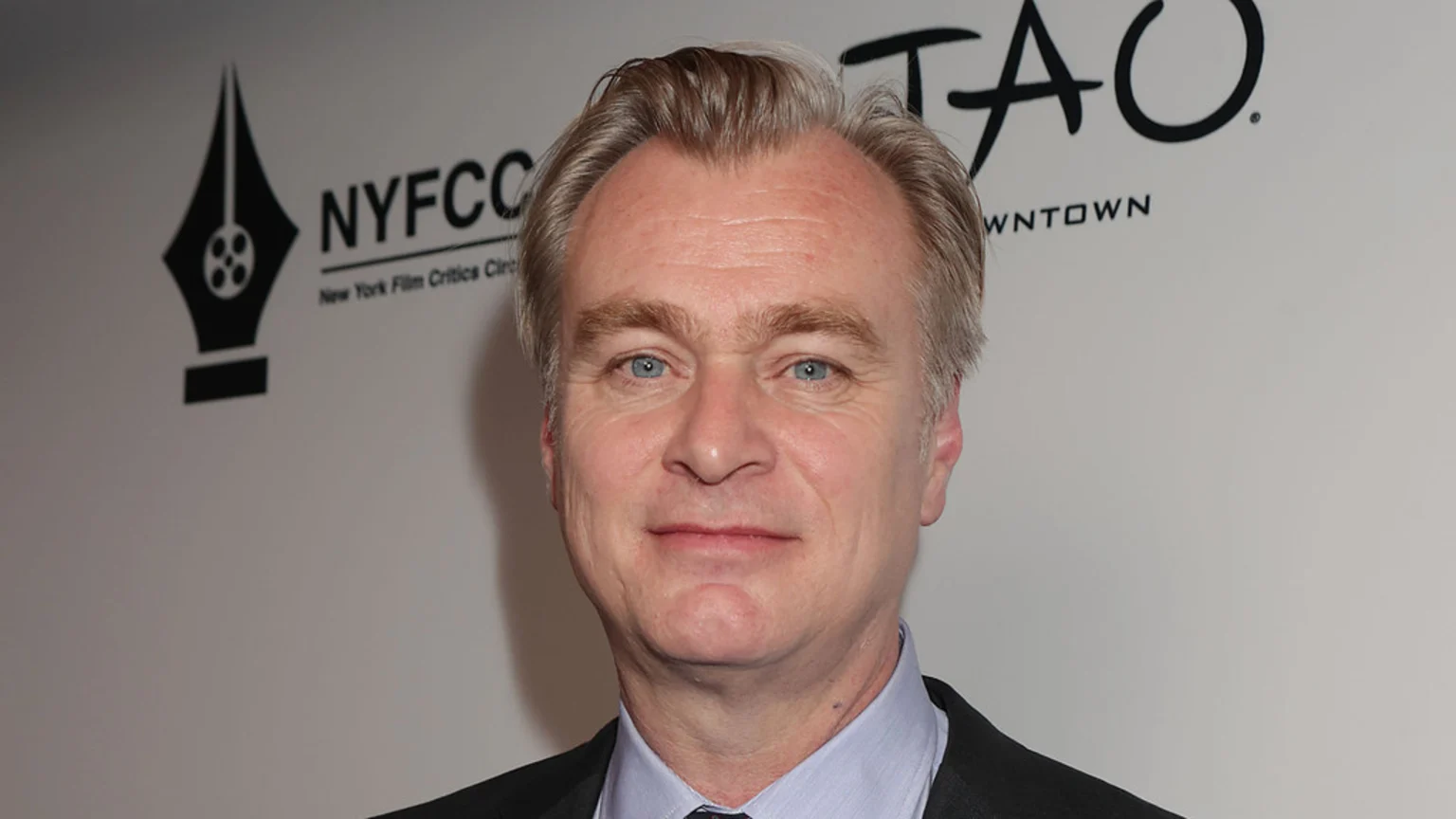 Christopher Nolan, a fair-skinned man with light hair and blue eyes, smiles faintly while wearing a suit and tie, with "NYFCC" and "TAO" logos visible in the background.