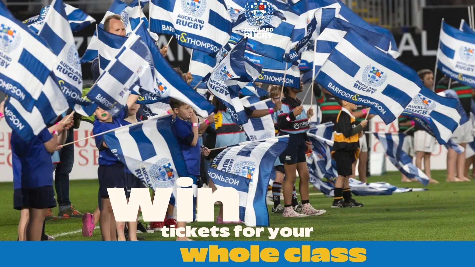Many children and some adults on a green field wave blue and white striped flags displaying "Auckland Rugby" and "Barfoot & Thompson."
