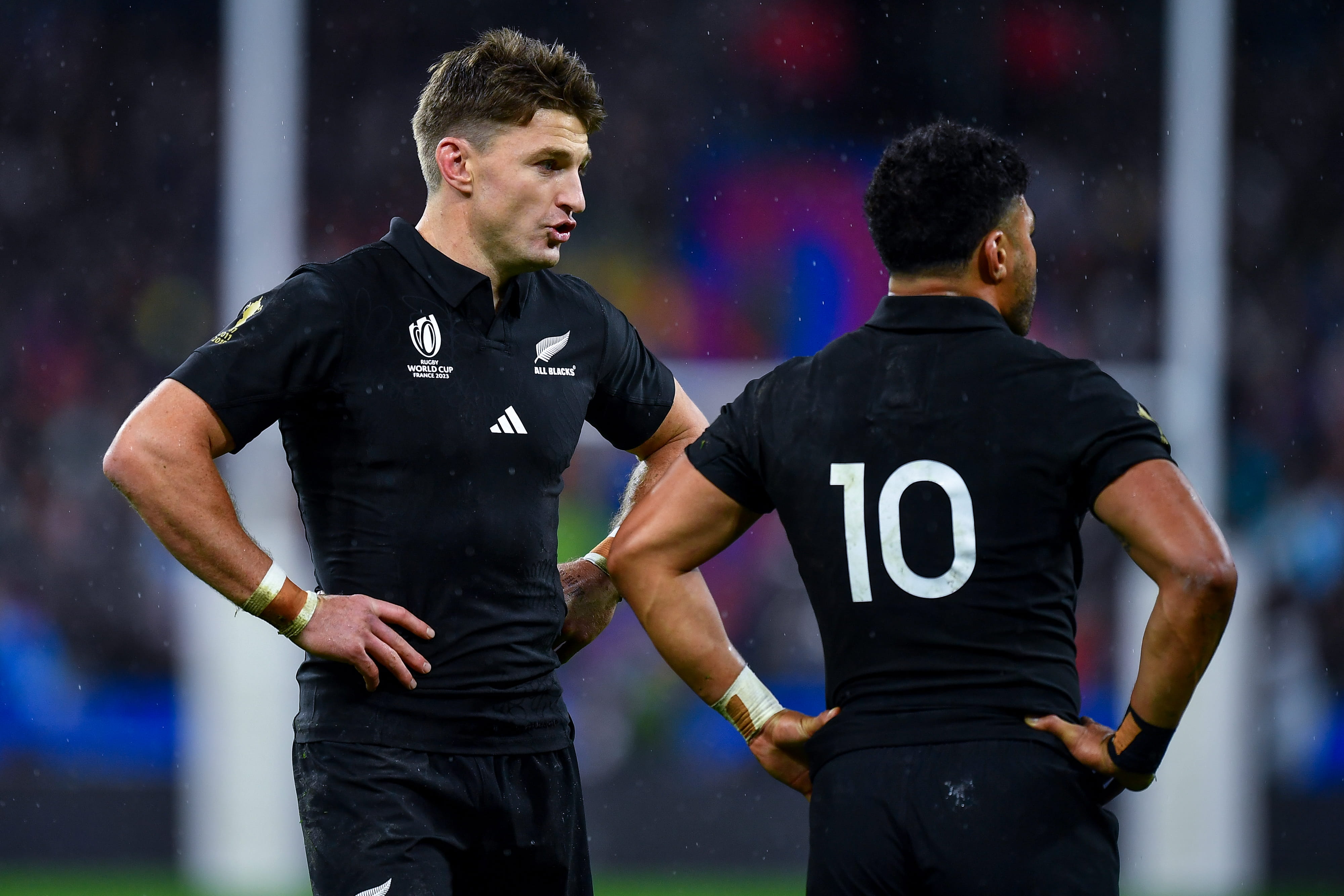Two All Blacks rugby players in black jerseys, one speaking to another whose number 10 jersey is visible from behind, on a wet field with stadium lights.