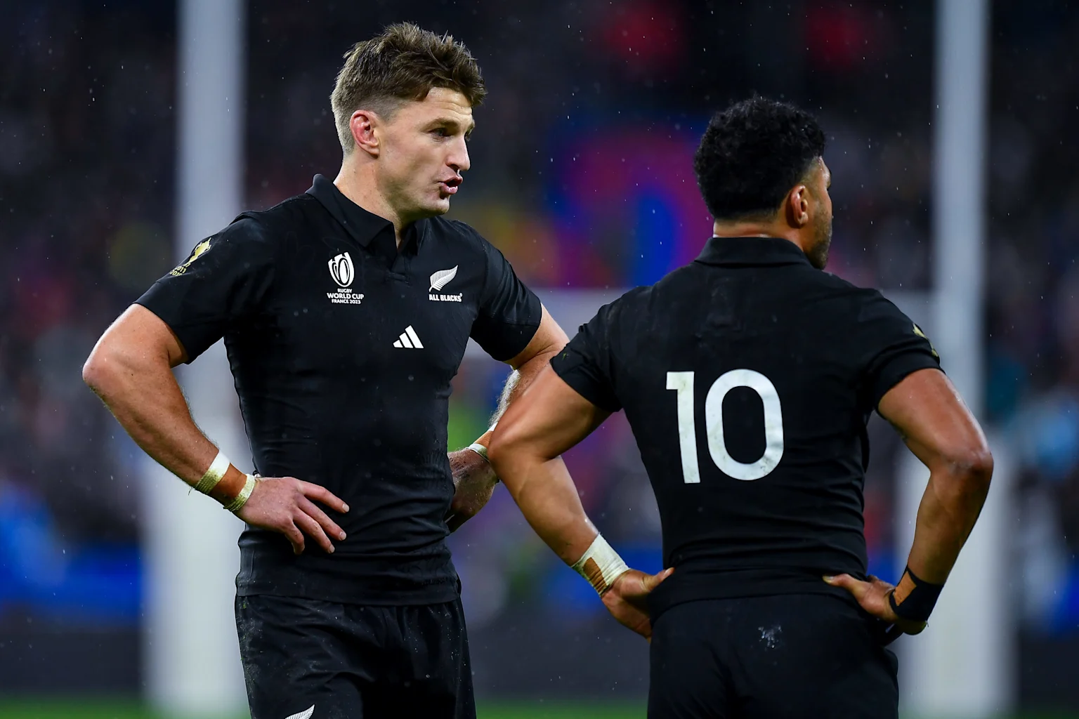 Two All Blacks rugby players in black jerseys, one speaking to another whose number 10 jersey is visible from behind, on a wet field with stadium lights.