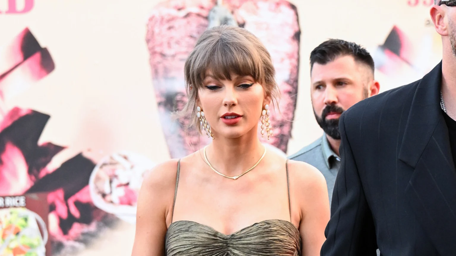 Taylor Swift in a gold strapless dress and dangling earrings, looking down, with men visible behind her against a blurry, light background with pink designs.