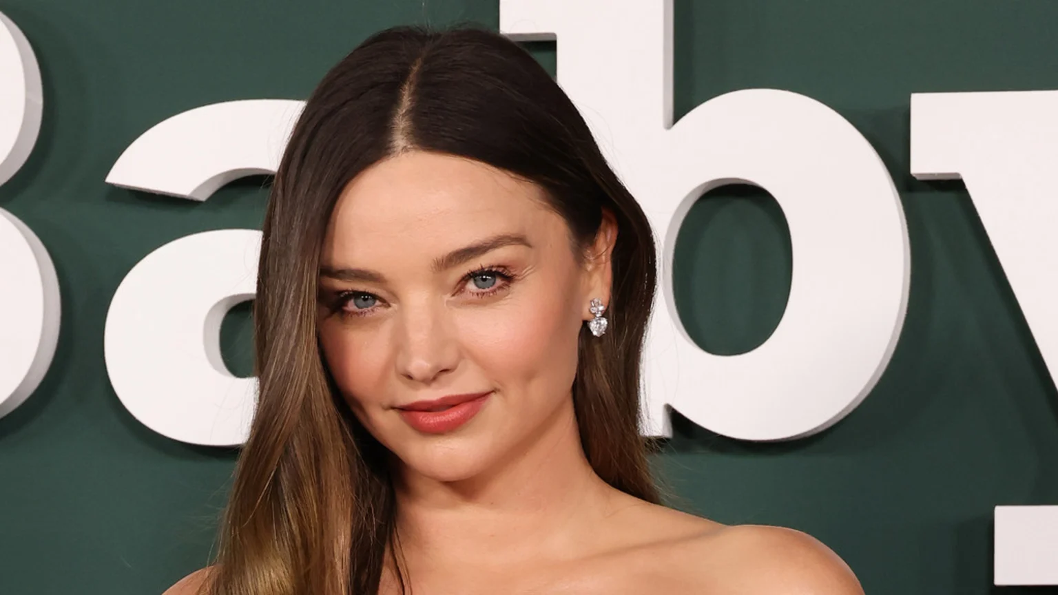 Close-up of Miranda Kerr with brown hair, diamond earrings, and a slight smile, against a dark green background with white letters forming "Baby".