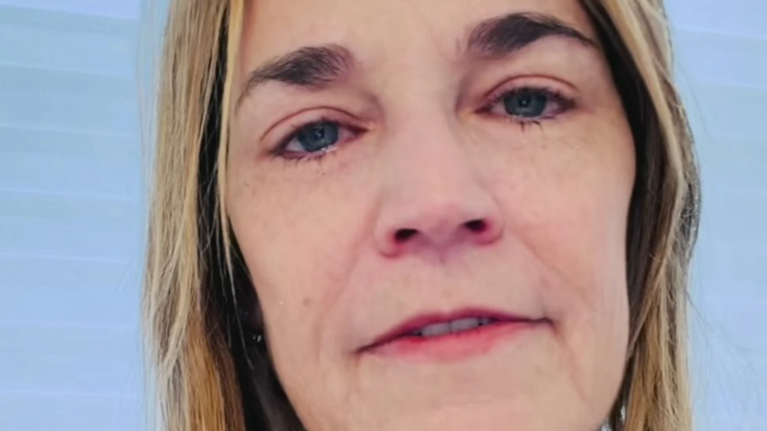 A close-up of a woman with blonde hair and blue eyes, appearing tearful or emotional, against a light blue background.