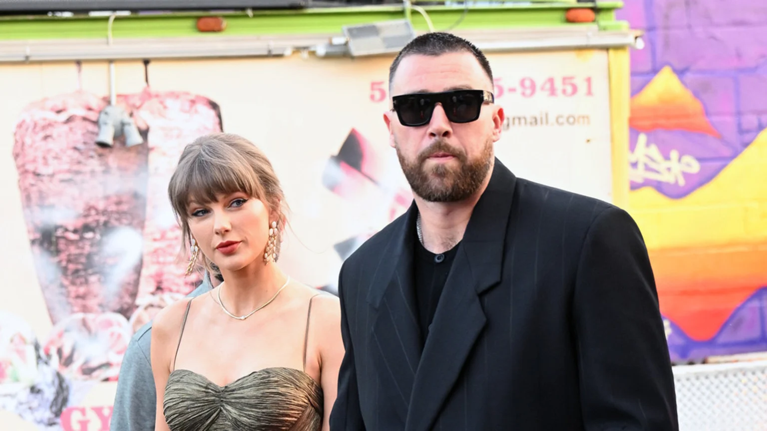 Taylor Swift in a strapless green dress stands next to Travis Kelce wearing sunglasses and a black suit, both looking forward.