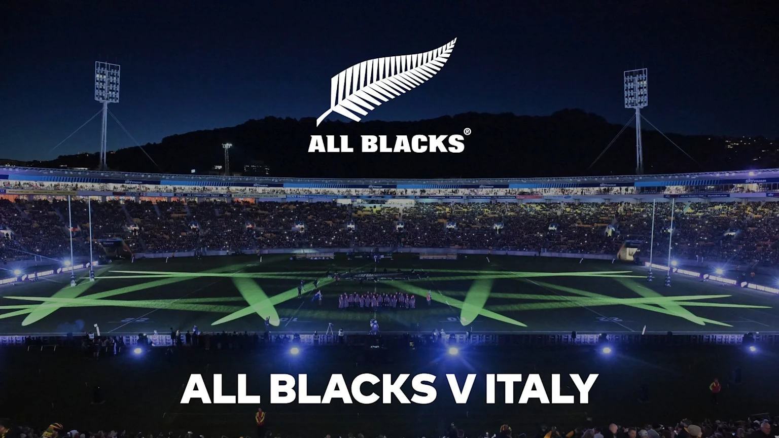 A night view of a rugby stadium filled with spectators, with the All Blacks logo and "ALL BLACKS V ITALY" text overlaying the image of the illuminated field where players are gathered.