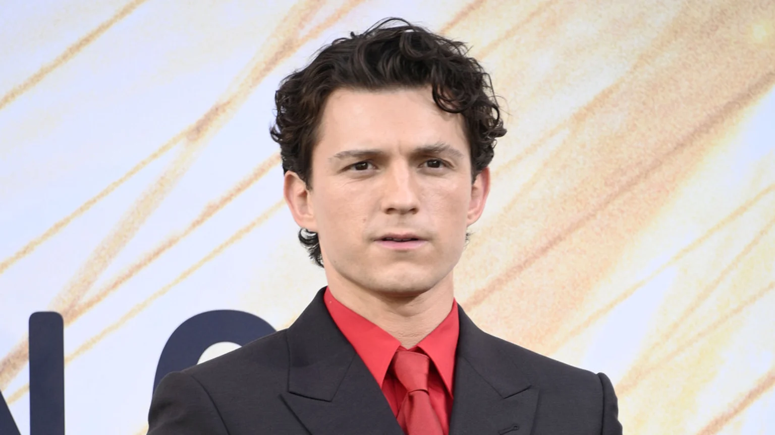 Actor Tom Holland looks at the camera, wearing a black suit, red shirt, and red tie, against a light background with abstract golden lines.