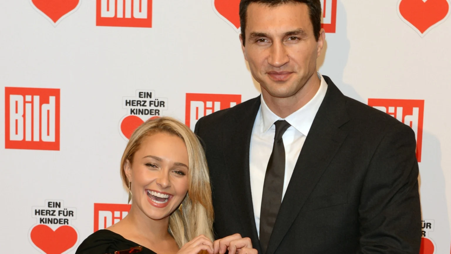 Wladimir Klitschko in a black suit smiles while standing next to Hayden Panettiere, who is laughing with her mouth open, against a white background with red "Bild" and "Ein Herz für Kinder" logos.