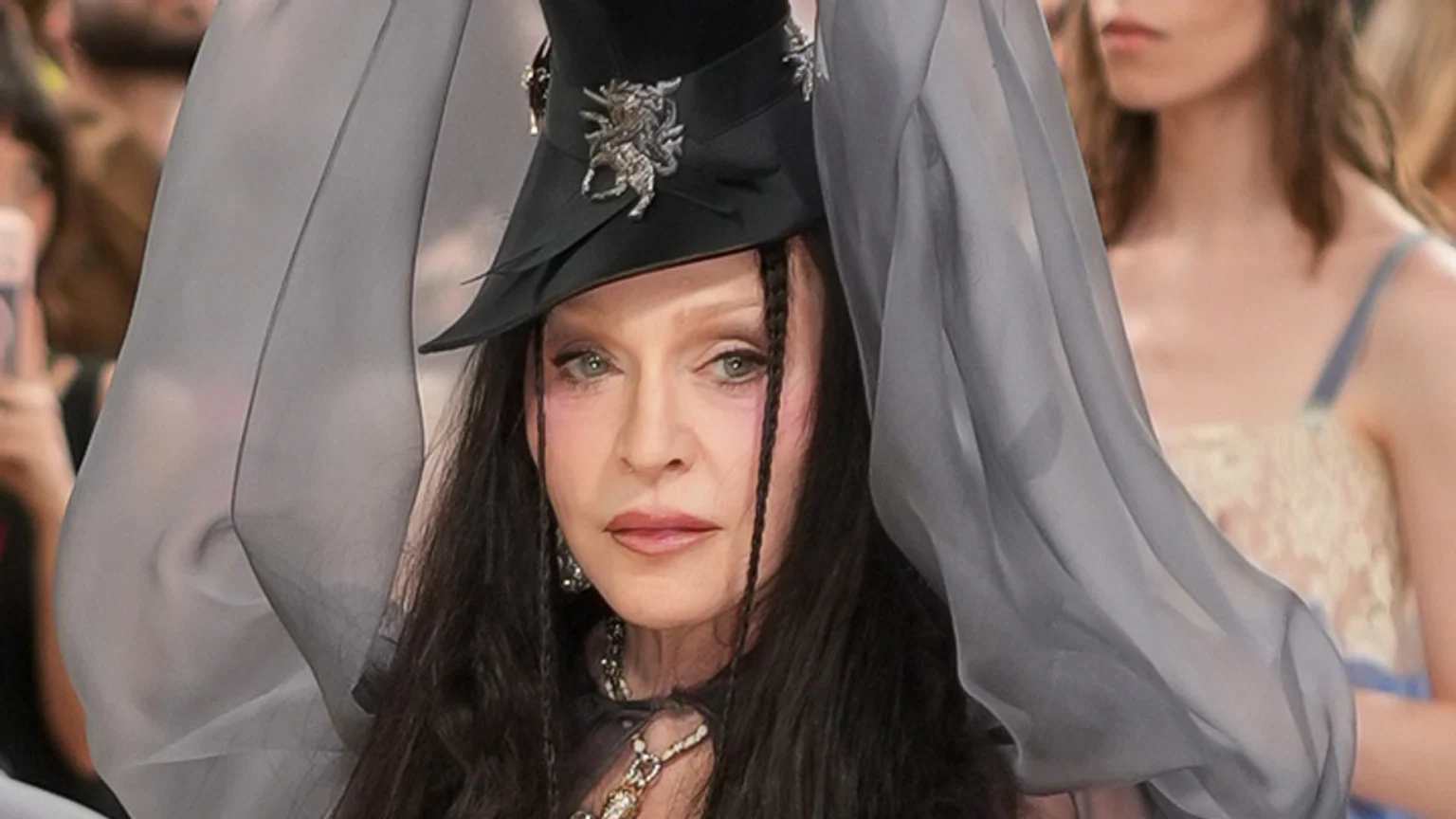 Close-up of Madonna with dark hair and prominent eye makeup, wearing a black hat with a silver ornament and sheer grey fabric framing her face.