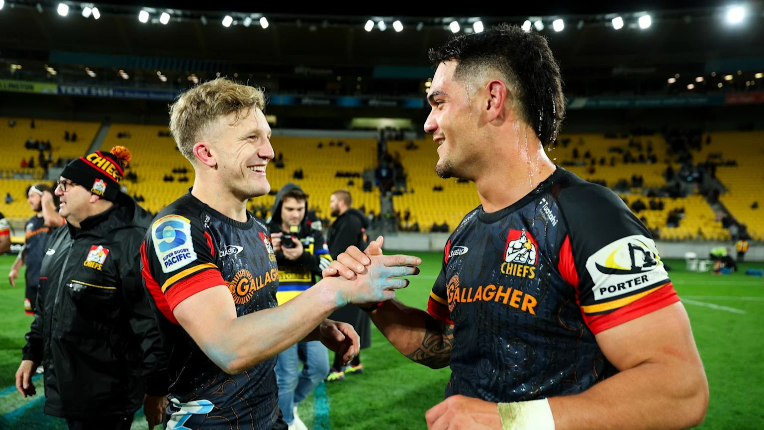 Two smiling rugby players in Chiefs jerseys shake hands on a field at night, one with blue on his hand, with stadium seats visible in the background.