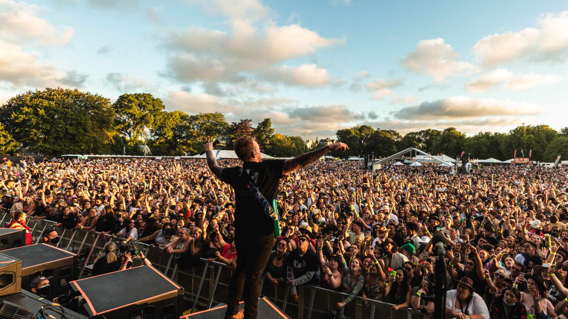 A musician on stage with arms outstretched, facing a massive crowd of people at an outdoor music festival, many with hands raised, under a cloudy sky.