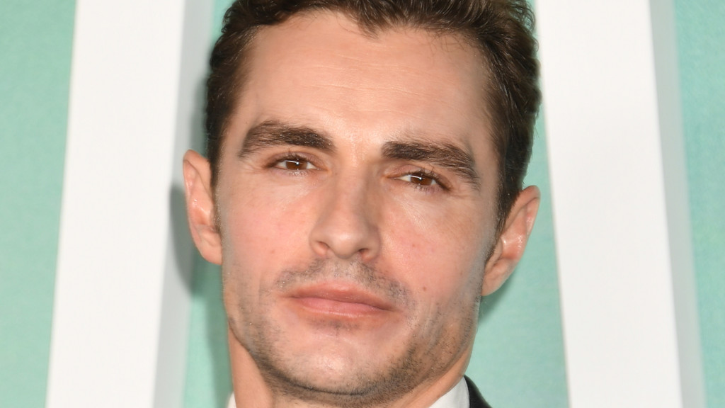 Close-up of actor Dave Franco looking directly at the camera with a neutral expression, against a blurred background of light green and white stripes.