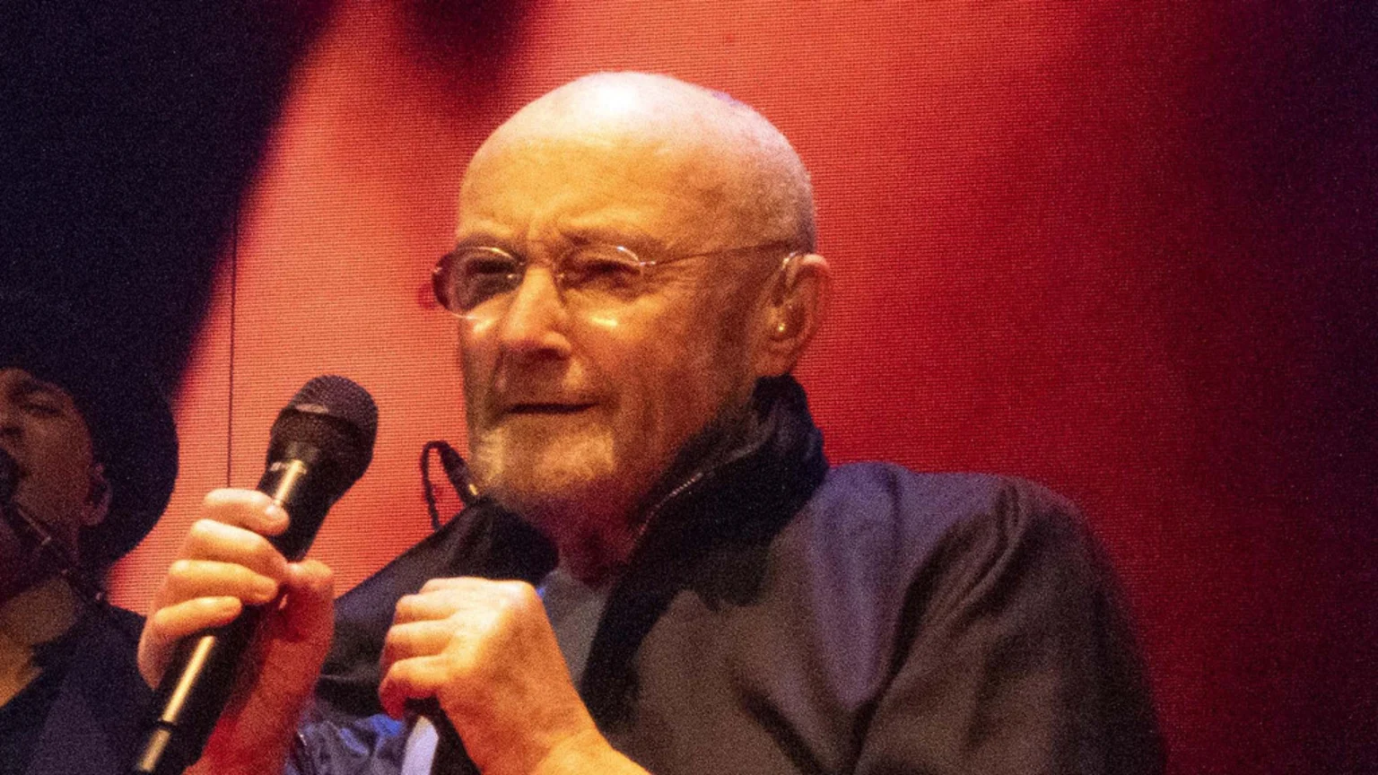 Close-up of Phil Collins, a bald man with glasses and a beard, holding a microphone and looking forward with a reddish background.