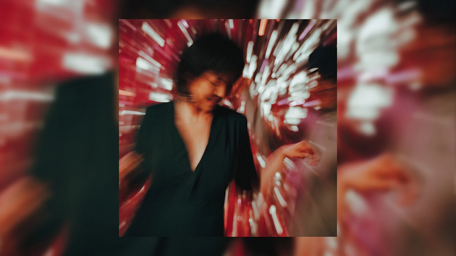 A person with dark hair, wearing a dark V-neck top, is captured in motion blur while dancing against a vibrant background of radial red and pink light streaks.
