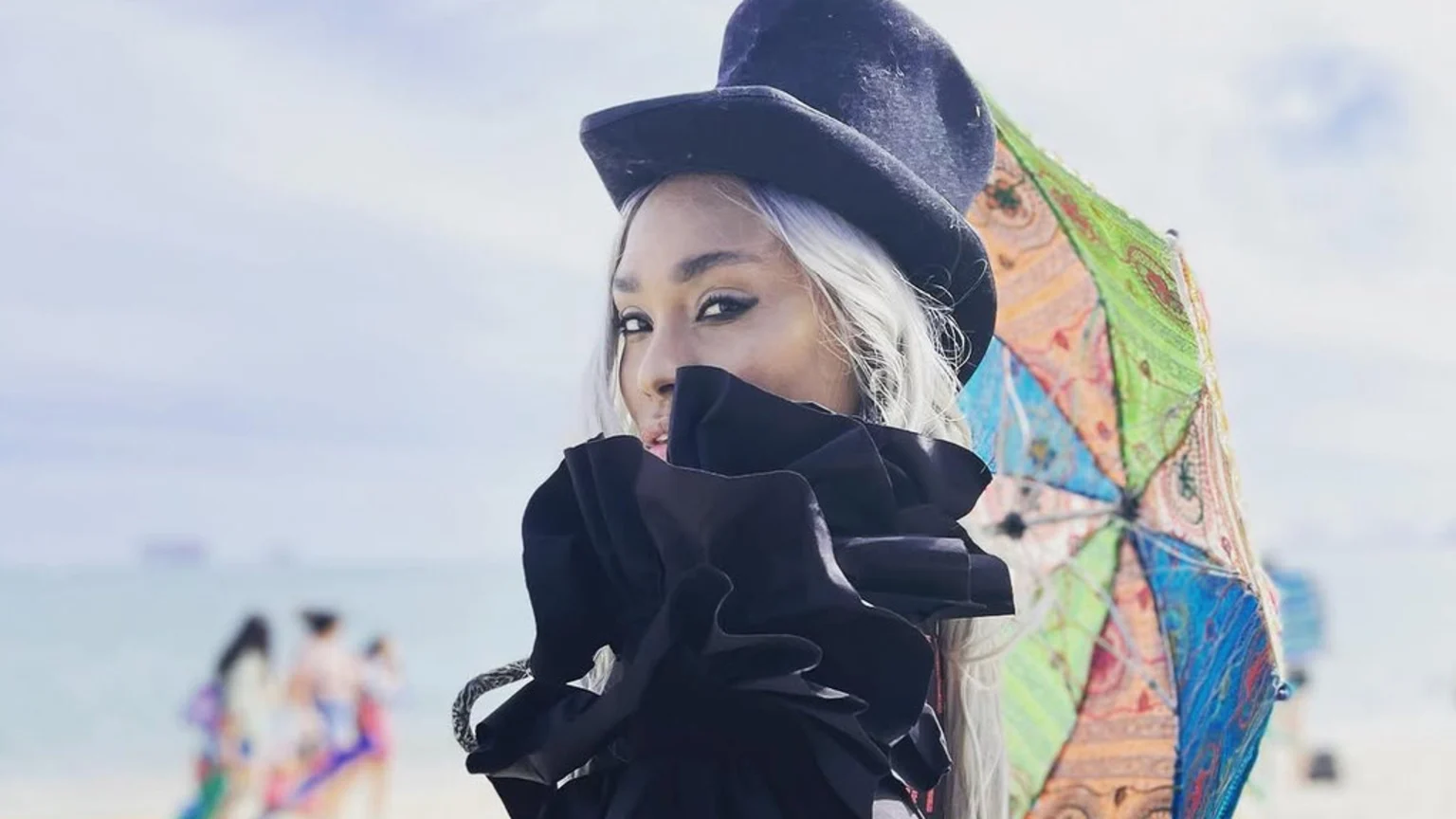 A person with platinum blonde hair, wearing a black top hat and a black ruffled garment over their mouth, looks at the camera on a bright beach with a colorful patterned umbrella to the right.