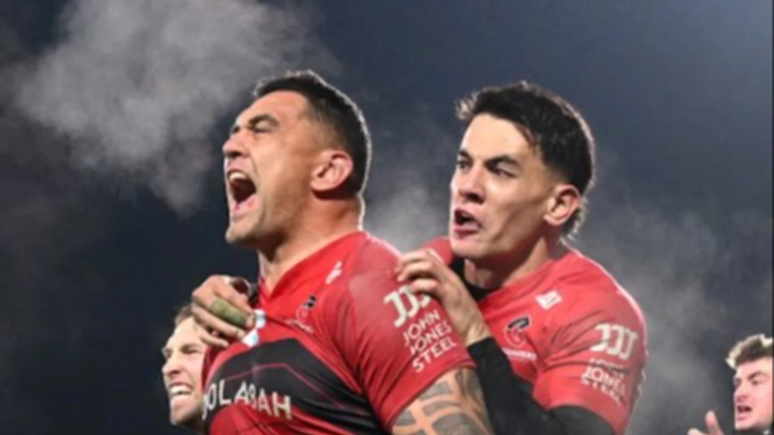 Two male rugby players in red and black jerseys celebrate with intense expressions, one yelling with visible breath in cold air, the other cheering with an arm around him.