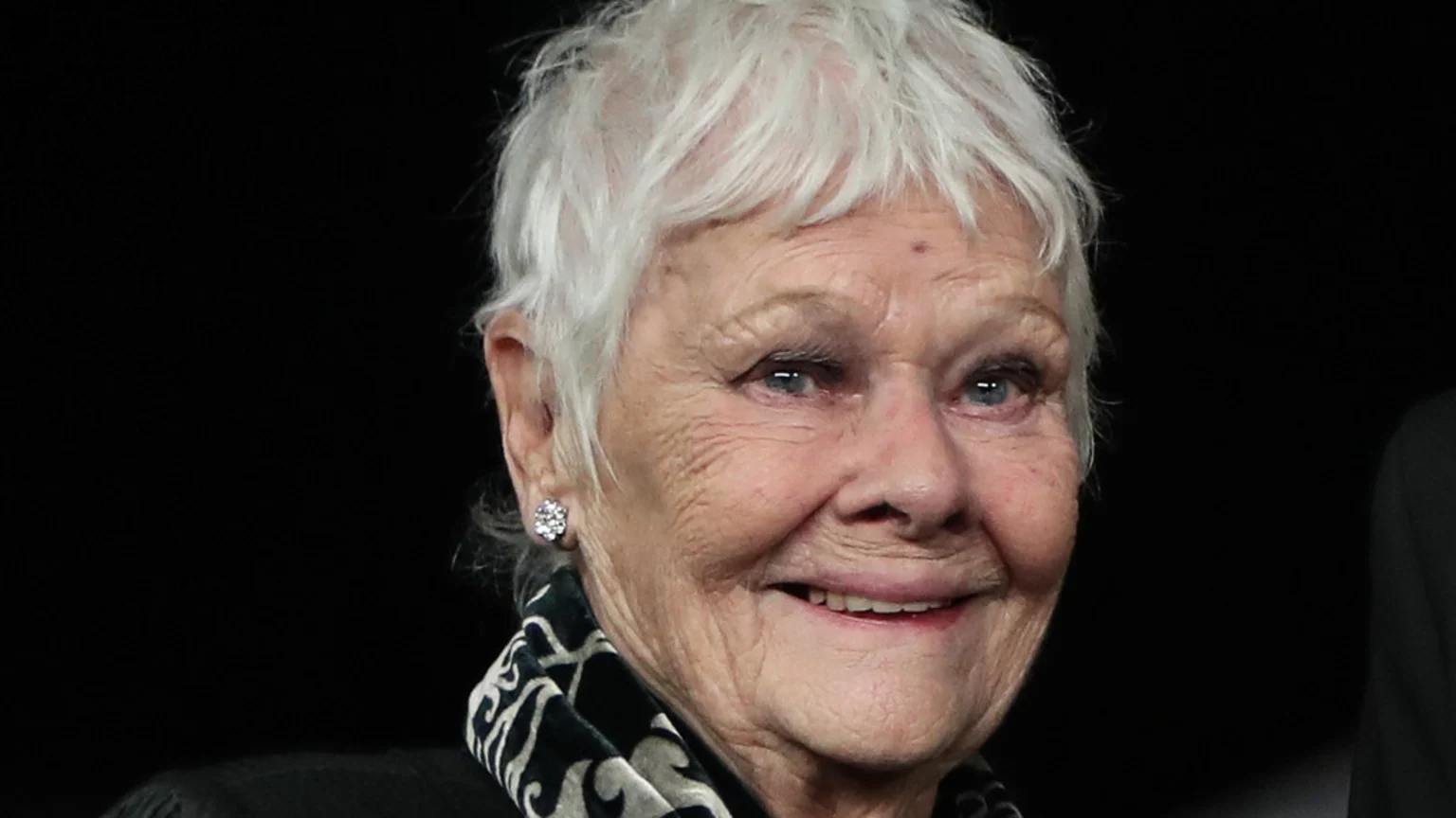 Close-up of Judi Dench, an elderly woman with short white hair and blue eyes, smiling and wearing a diamond earring and patterned scarf against a dark background.