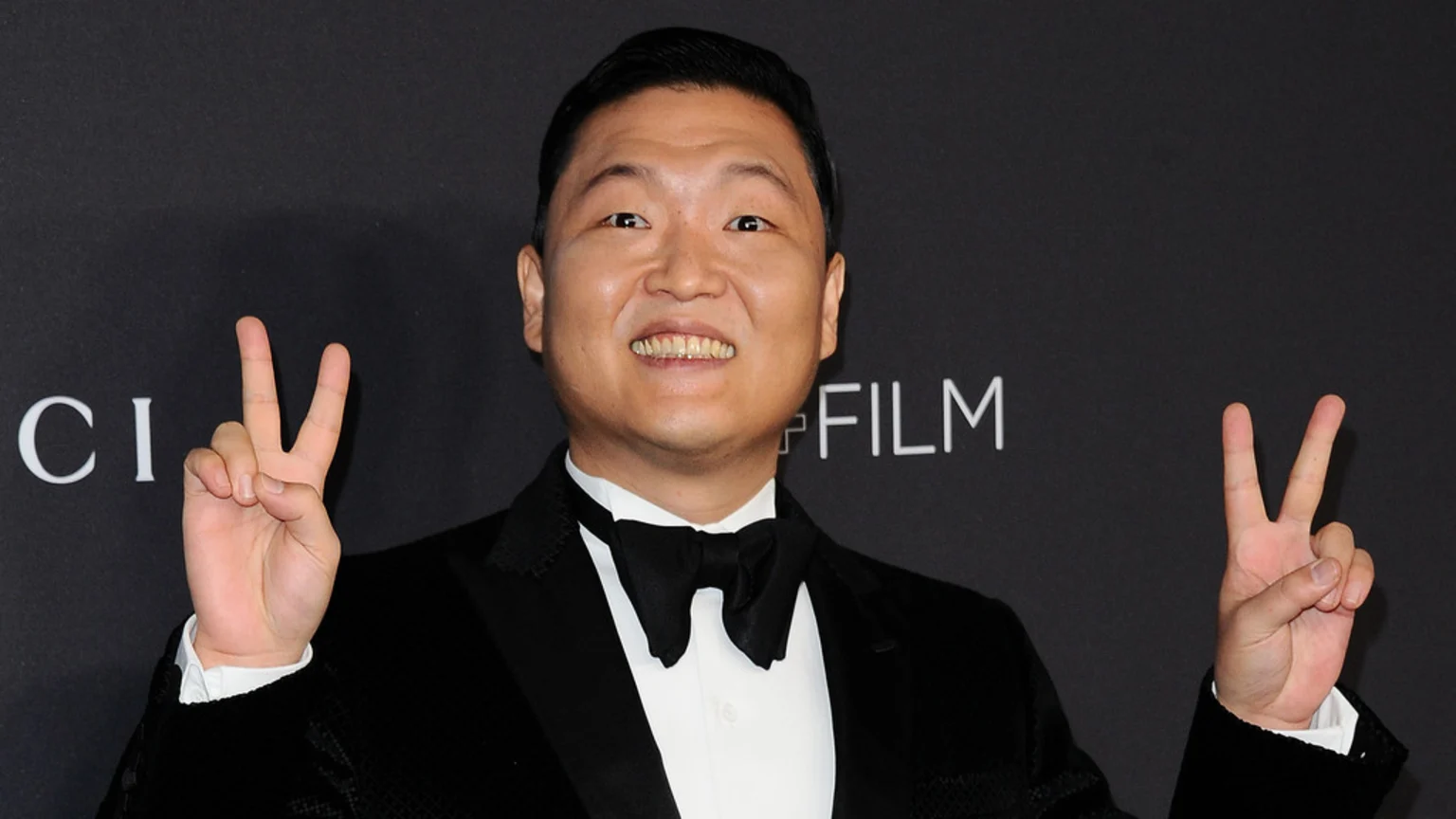 PSY, a man in a tuxedo, smiles and makes peace signs with both hands against a dark background with partially visible white text "GUCCI" and "FILM".