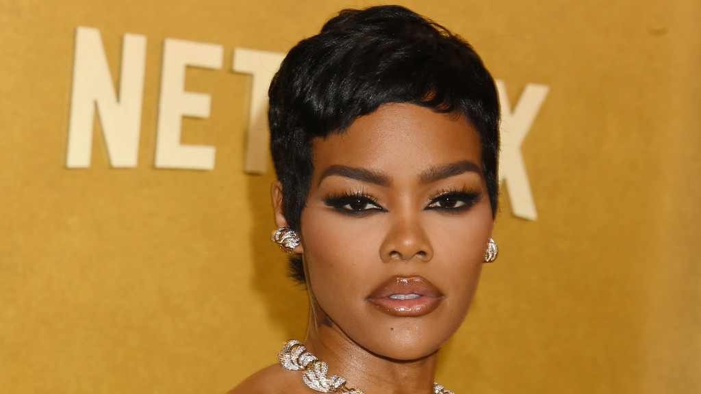 Close-up of Teyana Taylor with short black hair, dramatic eye makeup, glossy lips, and elaborate diamond earrings and necklace, against a blurry golden background with "NETFLIX" letters.