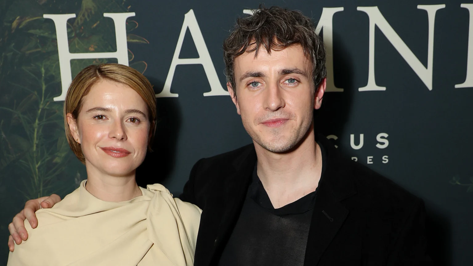 Paul Mescal with his arm around Jessie Buckley's shoulder, smiling at the camera in front of a "HAMNET" backdrop.