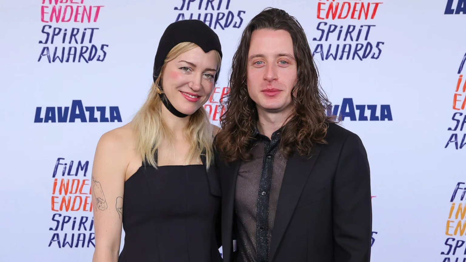 Rory Culkin and Kate Arizmendi pose together on a white step-and-repeat banner at the Independent Spirit Awards.